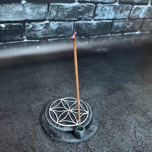 May include: A black incense holder with a white flower of life design. A brown incense stick is burning in the holder.