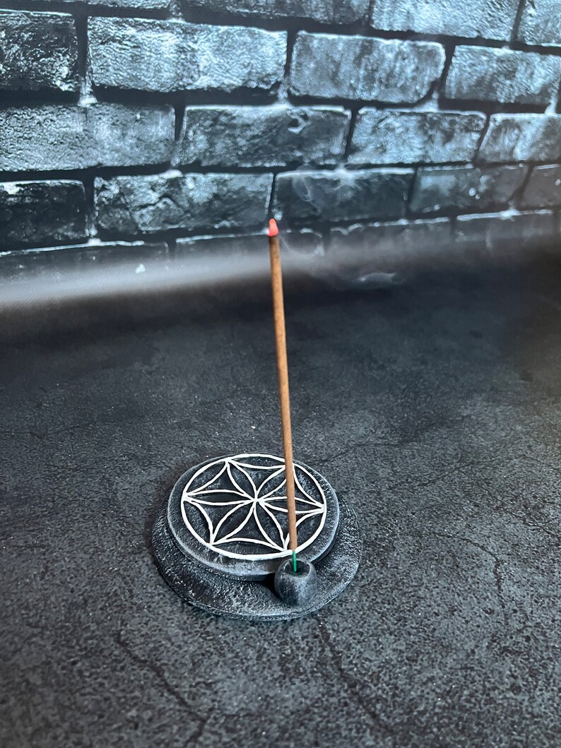May include: A black incense holder with a white flower of life design. A brown incense stick is burning in the holder.