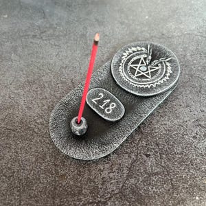 Puede incluir: Un porta incienso de forma ovalada con un acabado gris oscuro texturizado. Presenta un emblema circular con un pentagrama y figuras de dragones, una pequeña placa ovalada en relieve con el número 218 y un soporte tipo cuenta con una varilla de incienso roja.