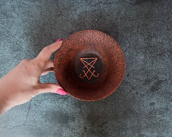 May include: A small, round, copper-colored bowl with a black sigil in the center. The bowl is slightly textured and has a metallic sheen.