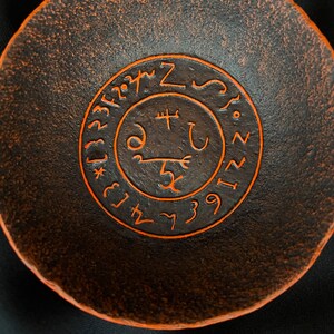 K&ouml;nnte beinhalten: Eine dunkelbraune Keramikschale mit einem orangefarbenen Sigil, das auf der Innenseite eingraviert ist. Das Sigil zeigt einen Kreis mit einem Kreuz im Inneren und einer Reihe von Symbolen um den Au&szlig;enrand.