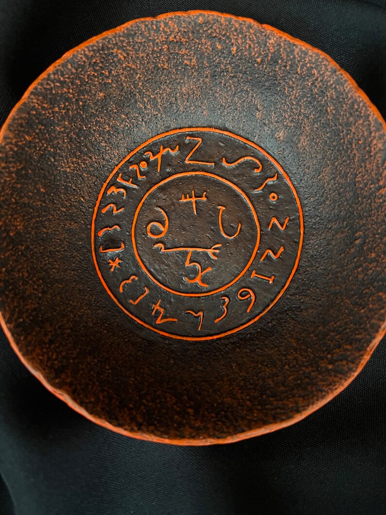 K&ouml;nnte beinhalten: Eine dunkelbraune Keramikschale mit einem orangefarbenen Sigil, das auf der Innenseite eingraviert ist. Das Sigil zeigt einen Kreis mit einem Kreuz im Inneren und einer Reihe von Symbolen um den Au&szlig;enrand.