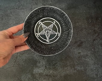 Pode incluir: Um descanso de copo de cerâmica preto com um pentagrama branco e uma cabeça de cabra dentro. O pentagrama tem o número 666 inscrito em cada ponto.