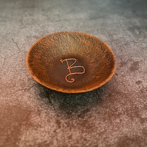 Op de afbeelding: Een klein, rond, bruin schaaltje met een getextureerd oppervlak. Het schaaltje heeft een enkele letter "B" gegraveerd in de bodem.