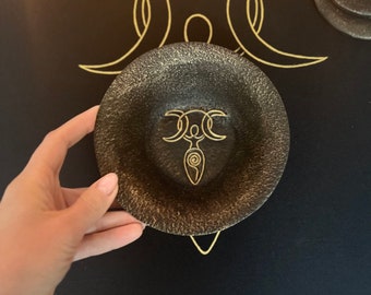 May include: A black bowl with a gold triple moon symbol and a stylised female figure inside the moon. The bowl is on a black background with a gold triple moon design.