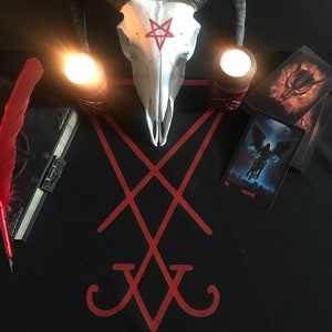 Pode incluir: Um pano preto com um símbolo de pentagrama vermelho e um crânio de carneiro branco com um pentagrama vermelho na testa. Duas velas acesas estão de cada lado do crânio. Uma pena vermelha e um livro fechado estão no lado esquerdo da imagem. Duas cartas de tarô estão no lado direito da imagem. A carta superior é uma imagem de fogo de um demônio e a carta inferior é uma imagem em preto e branco de uma figura alada com a palavra "LUCIFER" impressa nela.