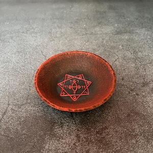 May include: A small, round, shallow bowl with a textured, matte finish. The exterior and rim are a vibrant, mottled red, while the interior deepens into a dark charcoal black. Centered in the bowl is a red, seven-pointed star with symbols.