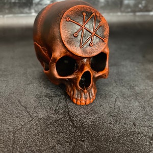 Könnte beinhalten: Ein schwarzer und orangefarbener Keramikschädel mit einem kreisförmigen Symbol auf der Stirn. Das Symbol ist ein Stern mit acht Zacken und einem Kreis darum.