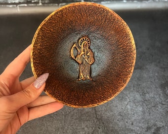 May include: A small, round, brown ceramic dish with a raised design of a figure in a hooded robe. The dish has a textured surface and a dark brown glaze.