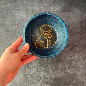 May include: A small, round, blue bowl with a gold-colored Egyptian hieroglyphic design. The bowl has a textured surface and a turquoise rim. The hieroglyphic design is of a bird with a sun disc above its head.