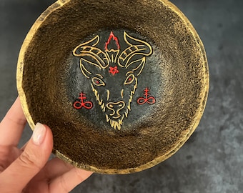 May include: A gold-coloured bowl with a black interior featuring a black and red goat's head with horns and a pentagram. The bowl is decorated with a red flame above the goat's head and two red upside-down crosses.