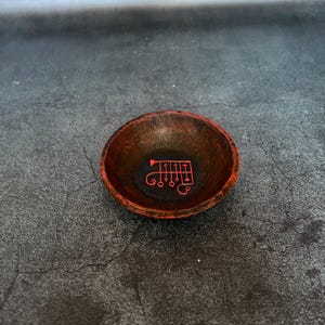 Puede incluir: Un cuenco pequeño, redondo y poco profundo con un acabado texturizado de color marrón rojizo oscuro. El interior del cuenco presenta un sigilo o símbolo rojo distintivo y centrado que consiste en líneas verticales con terminales circulares.