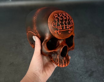 Può includere: Un teschio in ceramica nero e arancione con un sigillo rosso sulla fronte. Il teschio è tenuto in una mano.