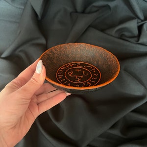K&ouml;nnte beinhalten: Eine schwarze Schale mit orangefarbenem Rand und einem kreisf&ouml;rmigen Design aus Symbolen und Zahlen. Die Schale ist aus einem strukturierten Material gefertigt.