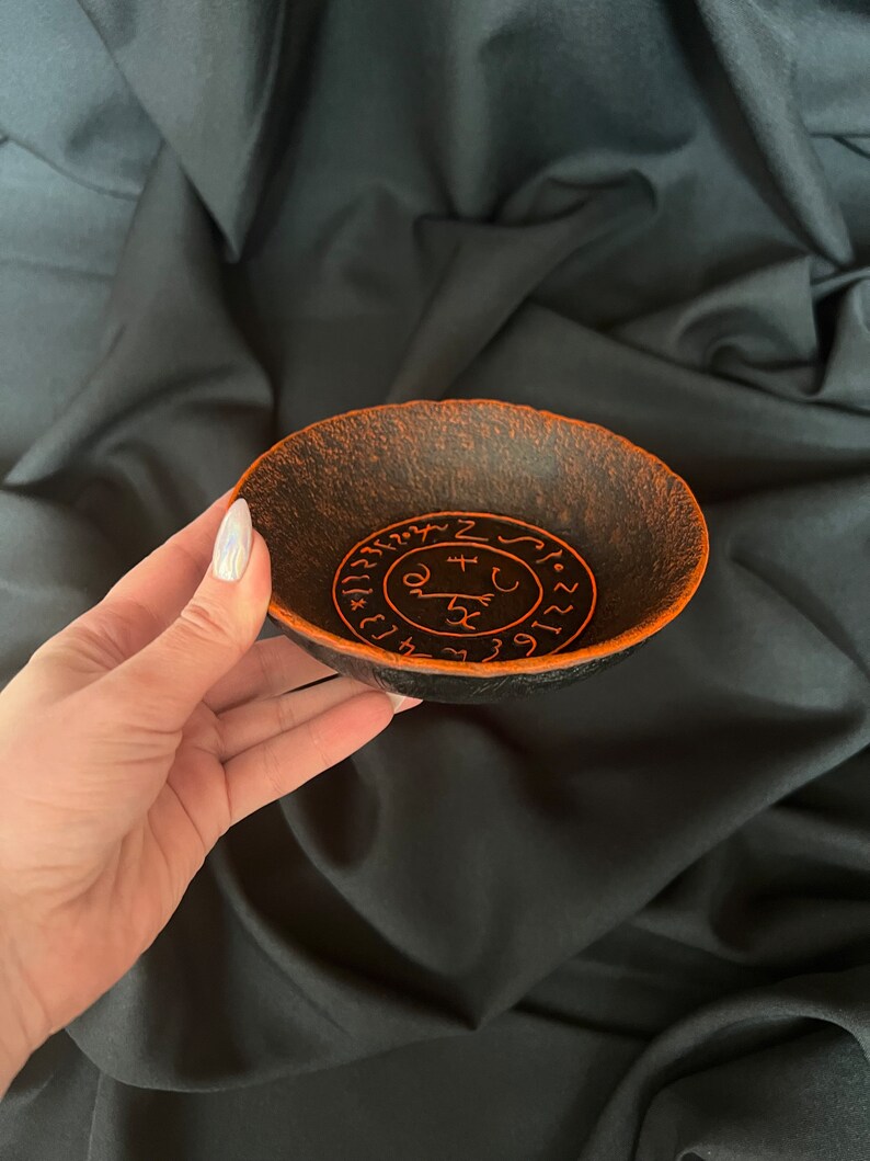 K&ouml;nnte beinhalten: Eine schwarze Schale mit orangefarbenem Rand und einem kreisf&ouml;rmigen Design aus Symbolen und Zahlen. Die Schale ist aus einem strukturierten Material gefertigt.