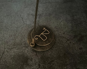May include: A gold metal incense holder with a round top and a single incense stick burning. The holder has the letter "D" engraved on it.