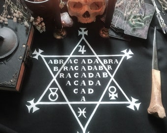 May include: A black cloth with a white pentagram and the words "ABRACADABRA" inscribed inside. The pentagram has a number "24" at the top and symbols at each point. A skull, candles, and other occult items are arranged around the cloth.