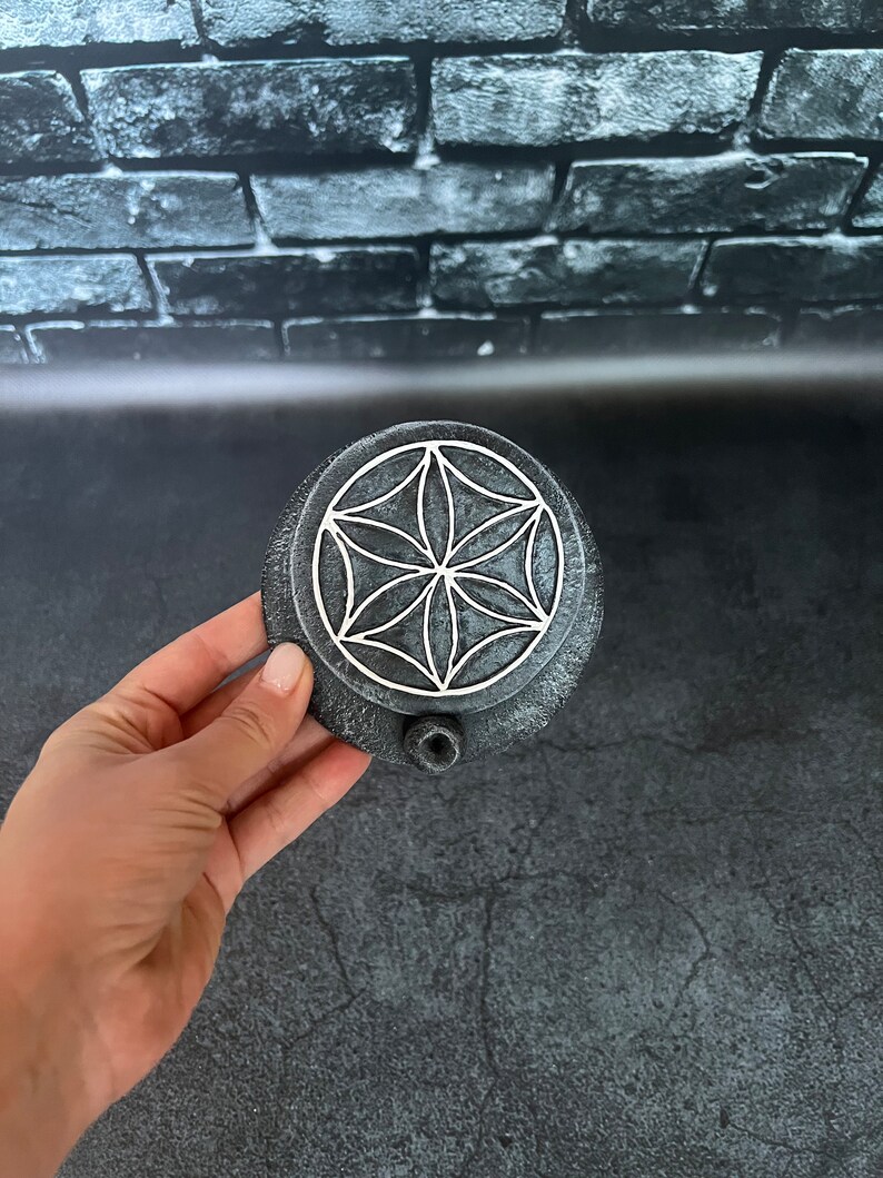 May include: A round, gray stone incense burner with a white geometric design. The burner has a small hole on the side for incense sticks.