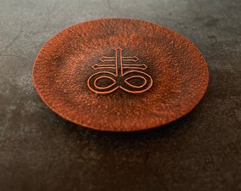 May include: A copper-colored metal coaster with a raised, black outline of a symbol that resembles a cross with an infinity symbol at the bottom.