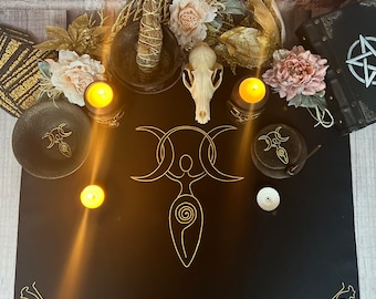 Puede incluir: Un paño de altar negro con un símbolo de triple luna dorado y una figura femenina estilizada. Hay velas, una calavera y un libro con un pentagrama en la portada.