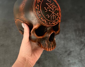 May include: A black and orange skull with a circular design on the forehead. The design features a pentagram with a symbol inside and is outlined in orange.