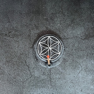 May include: A black circular incense holder with a white geometric design and a single incense stick burning in the center.