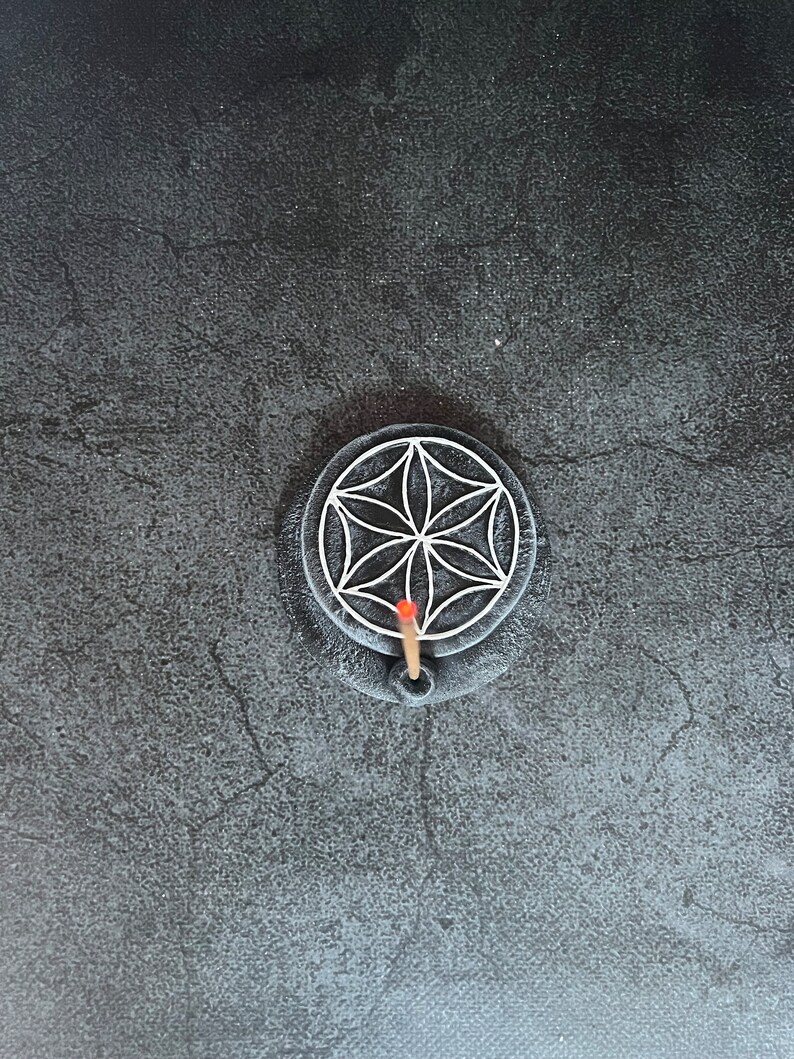 May include: A black circular incense holder with a white geometric design and a single incense stick burning in the center.