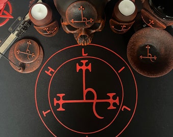 May include: A black skull with a red sigil on its forehead sits on a black surface with a red sigil circle. Two candles with red sigils are on the surface, along with a small bowl and a book.