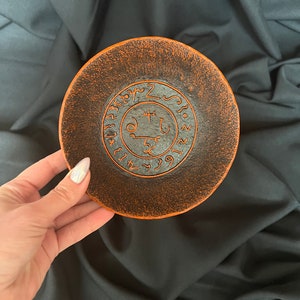K&ouml;nnte beinhalten: Eine kleine, runde, orangefarbene und schwarze Keramikschale mit einem schwarzen Sigil, das in der Mitte eingraviert ist. Das Sigil ist von einem Kreis aus schwarzem Text umgeben.