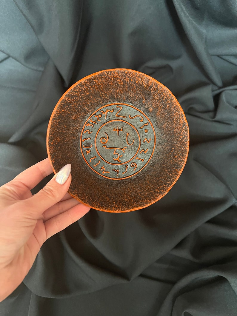 K&ouml;nnte beinhalten: Eine kleine, runde, orangefarbene und schwarze Keramikschale mit einem schwarzen Sigil, das in der Mitte eingraviert ist. Das Sigil ist von einem Kreis aus schwarzem Text umgeben.