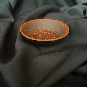 K&ouml;nnte beinhalten: Eine schwarze Schale mit einem orangefarbenen Design eines Kreises mit Symbolen darin. Die Schale befindet sich auf einem schwarzen Stoffhintergrund.