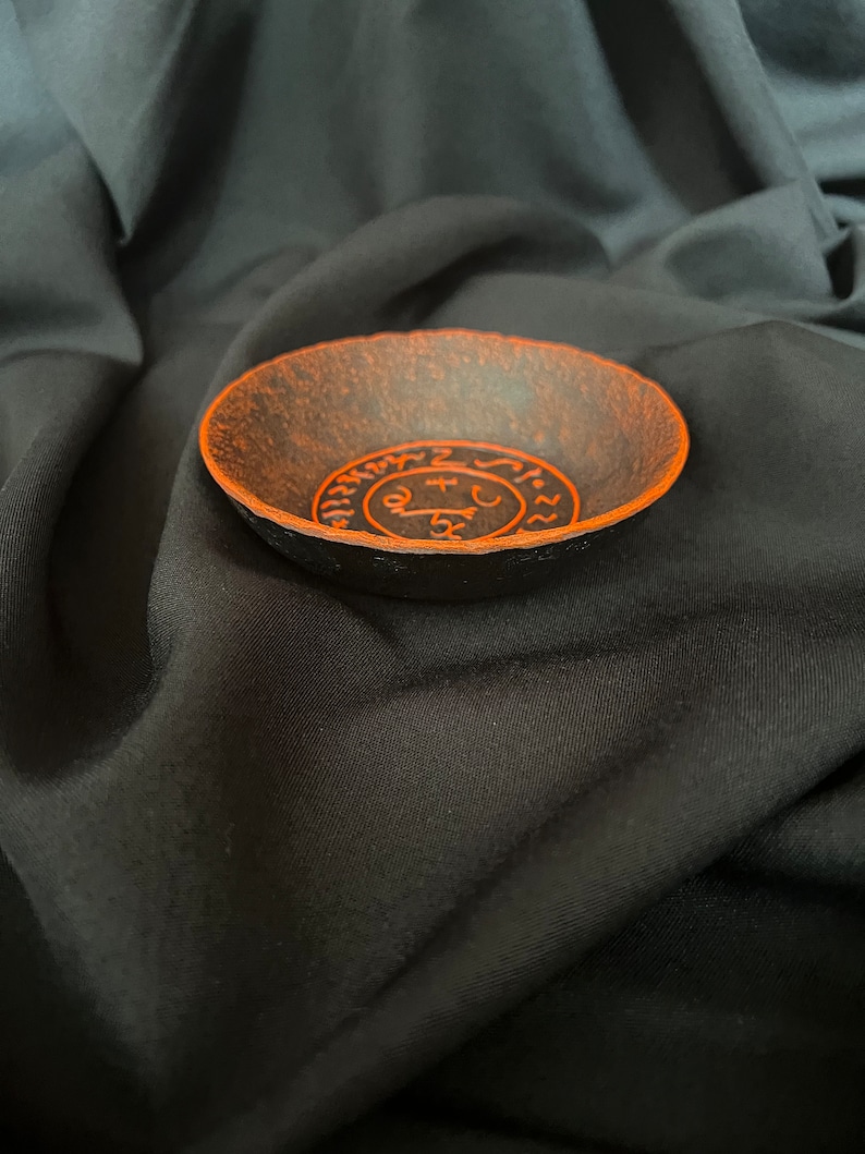 K&ouml;nnte beinhalten: Eine schwarze Schale mit einem orangefarbenen Design eines Kreises mit Symbolen darin. Die Schale befindet sich auf einem schwarzen Stoffhintergrund.