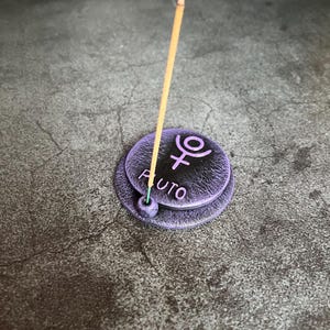 May include: Purple ceramic incense holder with the symbol for Pluto and the word "Pluto" inscribed on it. A wooden incense stick is burning in the holder.