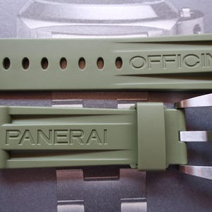 Puede incluir: Correa de reloj verde oliva con la palabra "OFFICINE" en relieve en la correa superior y "PANERAI" en la correa inferior. La correa tiene varios agujeros para ajustar y una hebilla plateada.