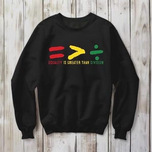 Op de afbeelding: Zwart sweatshirt met de tekst "EQUALITY IS GREATER THAN DIVISION". Het ontwerp toont een rood gelijkteken, een geel groter-dan-teken en een groen deelteken.