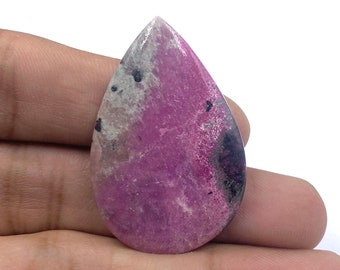 May include: A teardrop-shaped gemstone with a vibrant magenta hue, speckled with black and gray inclusions. The polished surface reflects light, showcasing the stone's natural patterns. The gemstone is held in a hand, highlighting its size and shape.