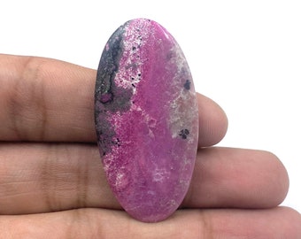 May include: An oval-shaped cabochon gemstone featuring a vibrant magenta and pink surface with contrasting dark gray and black mineral inclusions. The stone has a smooth, polished finish and displays a natural, mottled pattern across its entire face.