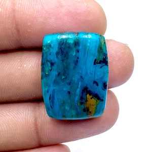 Op de afbeelding: Een gepolijste, rechthoekige edelsteen met een levendig blauw en turkoois kleurenschema. De steen heeft donkerblauwe en goudkleurige insluitsels, waardoor een uniek patroon ontstaat. De edelsteen wordt tussen vingers gehouden.