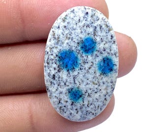 May include: Oval-shaped K2 stone, featuring a speckled white and gray base with vibrant blue spots. The polished gemstone is held in a hand, showcasing its natural patterns and colors. This unique stone is ideal for jewelry making or collecting.