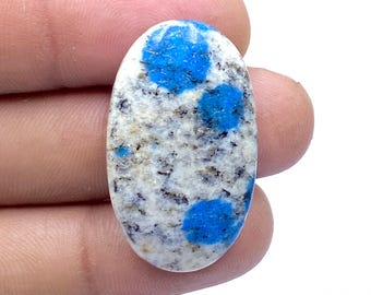 May include: Oval-shaped K2 stone with a white base speckled with black and vibrant blue spots. The polished gemstone is held in a hand, showcasing its natural patterns and colors. This stone is often used in jewelry.