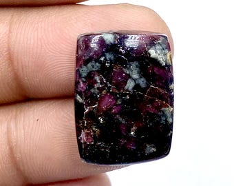 Puede incluir: Una gema rectangular con una mezcla de colores rojo oscuro, negro y gris. La piedra tiene una superficie pulida y un aspecto natural y texturizado. La gema se sostiene entre los dedos.