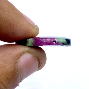 May include: A multi-coloured gemstone pendant with a hole drilled through it. The stone features a mix of colours, including deep red, green, and black. The pendant is held between two fingers against a white background.