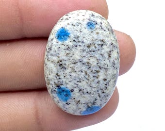 May include: Oval-shaped K2 stone cabochon. The stone features a speckled white and gray base with vibrant blue spots. The polished surface highlights the natural patterns and colors of the gemstone.
