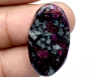 Puede incluir: Una piedra preciosa de forma ovalada con una mezcla de colores. La piedra presenta una base oscura, casi negra, salpicada de gris y tonos rojo-púrpura vibrantes. La superficie pulida refleja la luz.