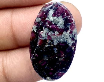 Puede incluir: Una piedra preciosa ovalada con una mezcla de colores borgoña oscuro, grises y negros. La piedra tiene una superficie pulida y un patrón único, con los colores mezclándose en un efecto veteado. La gema está sujeta en la mano de una persona.
