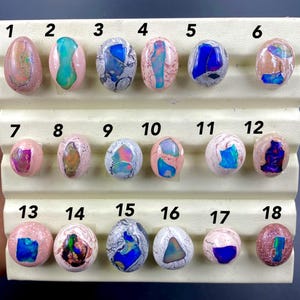 May include: A collection of eighteen oval-shaped gemstones, each with unique patterns and colors. The gemstones are set on a white display stand. Colors include pink, blue, white, and brown. Each stone is numbered from 1 to 18.