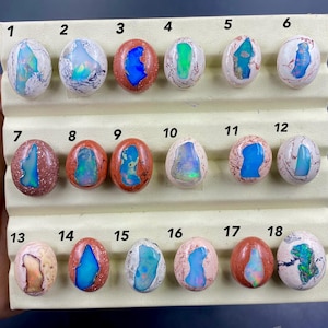 May include: Eighteen oval opals are displayed, each with distinct patterns and colours. The opals are set on a beige display. The opals feature a variety of colours, including blue, green, and orange, with some having a marbled appearance.