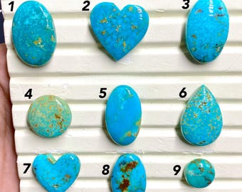 May include: A collection of nine turquoise gemstones in various shapes, including oval, heart, and round. The gemstones are a vibrant blue-green color with brown matrix patterns. The stones are displayed on a white surface.