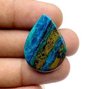 Könnte beinhalten: Ein tropfenförmiger Cabochon aus blauer und grüner Chrysocolla mit braunen und goldenen Einschlüssen. Der Stein hat eine glatte, polierte Oberfläche.