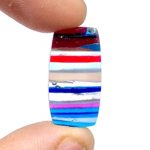 May include: A clear glass cabochon with a layered pattern of blue, pink, red, and white stripes. The cabochon is oval shaped and has a smooth, polished surface.